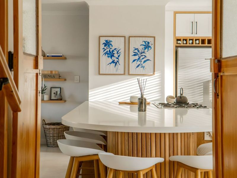A bright, modern kitchen featuring a curved wooden island with a white countertop, built-in cooktop, and minimalist floating shelving.