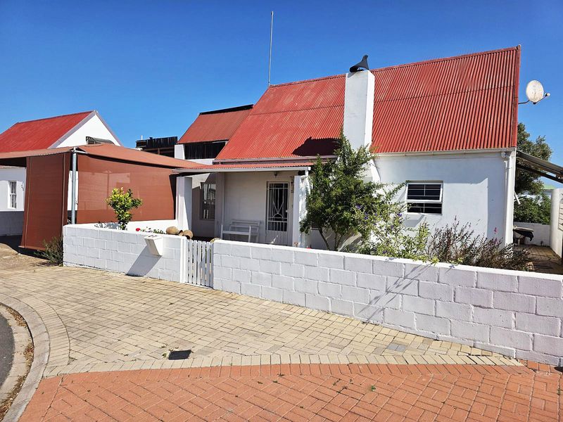 Front exterior view of a cottage-style house featuring a red corrugated metal roof, white masonry walls, and a paved driveway with a boundary wall.