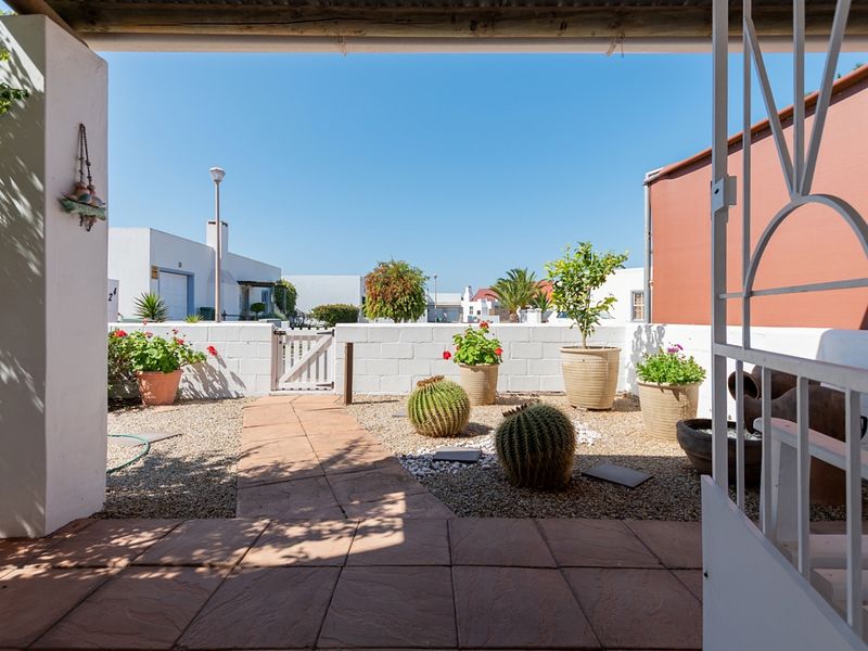 A covered patio area featuring tiled flooring, overlooking a landscaped courtyard with a paved walkway, gravel garden beds, and a secure boundary wall.
