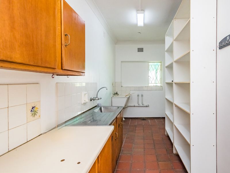 A functional scullery or laundry area featuring built-in wooden cabinetry, a stainless steel sink, utility basin, and durable terracotta floor tiles.