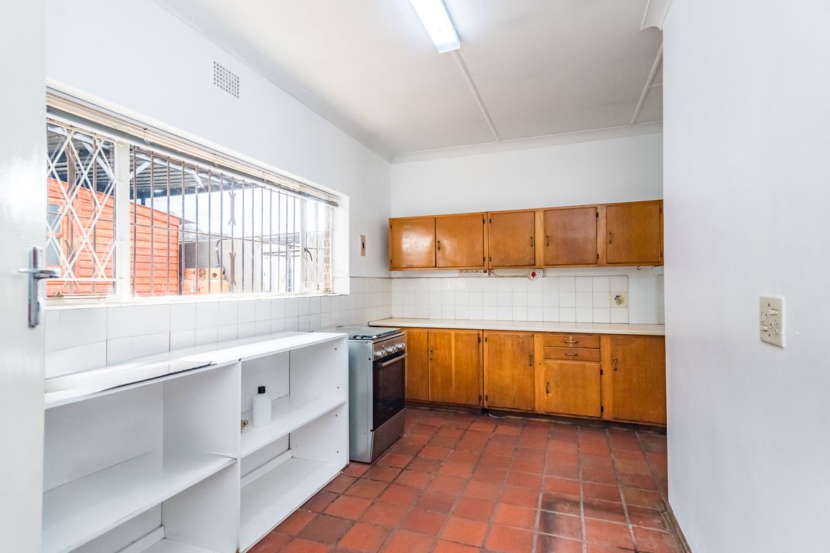 A functional kitchen featuring wooden cabinetry, a tiled backsplash, a freestanding stove, and durable terracotta floor tiles with security bars on the window.