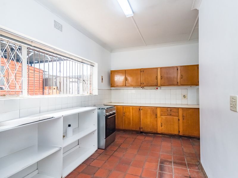 A functional kitchen featuring wooden cabinetry, a tiled backsplash, a freestanding stove, and durable terracotta floor tiles with security bars on the window.