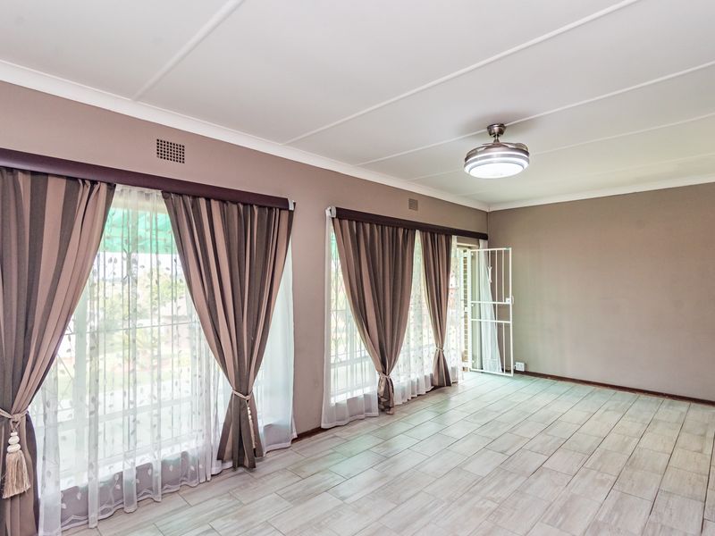 Spacious living area featuring neutral wall tones, light-colored tiled flooring, a ceiling fan, and large windows with security gates.