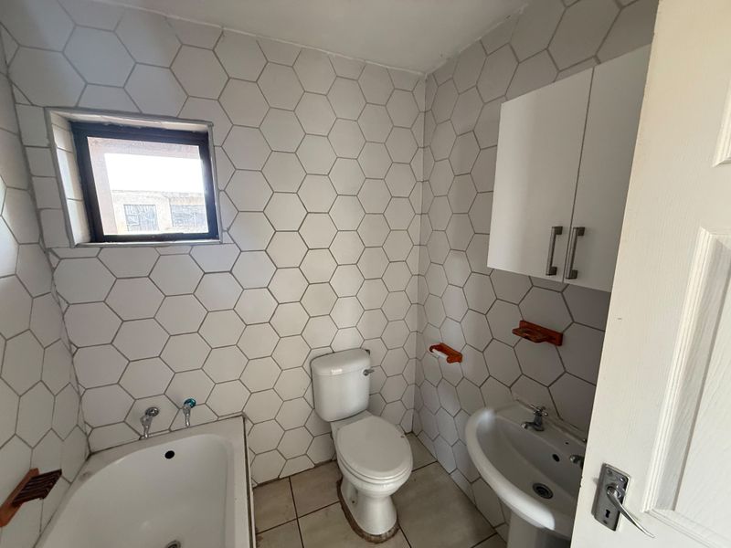 A functional bathroom featuring hexagonal wall tiling, a standard bathtub, pedestal basin, and wall-mounted storage cabinet.