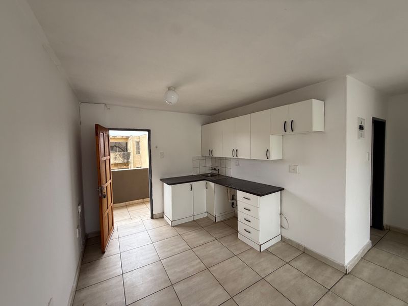 A compact, empty kitchen featuring white built-in cabinetry, dark laminate countertops, a stainless steel sink, and tiled flooring with direct access to a balcony.