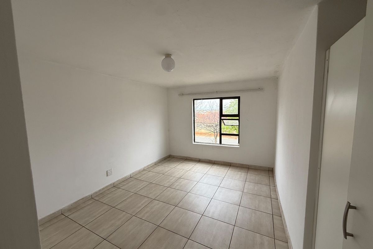 A clean, empty bedroom featuring neutral tiled flooring, a built-in cupboard, and a window providing natural light.