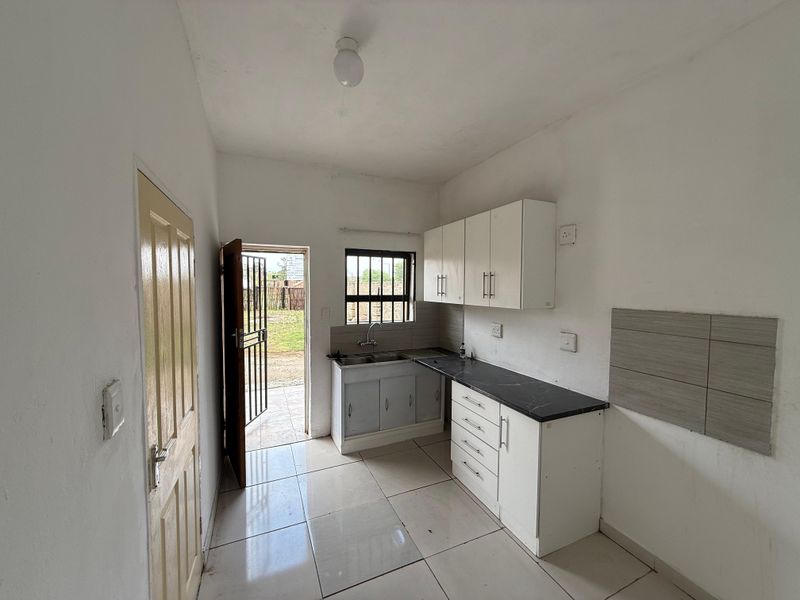 A compact, empty kitchen featuring white cabinetry, dark laminate countertops, tiled flooring, and direct access to an outdoor area through a security gate.