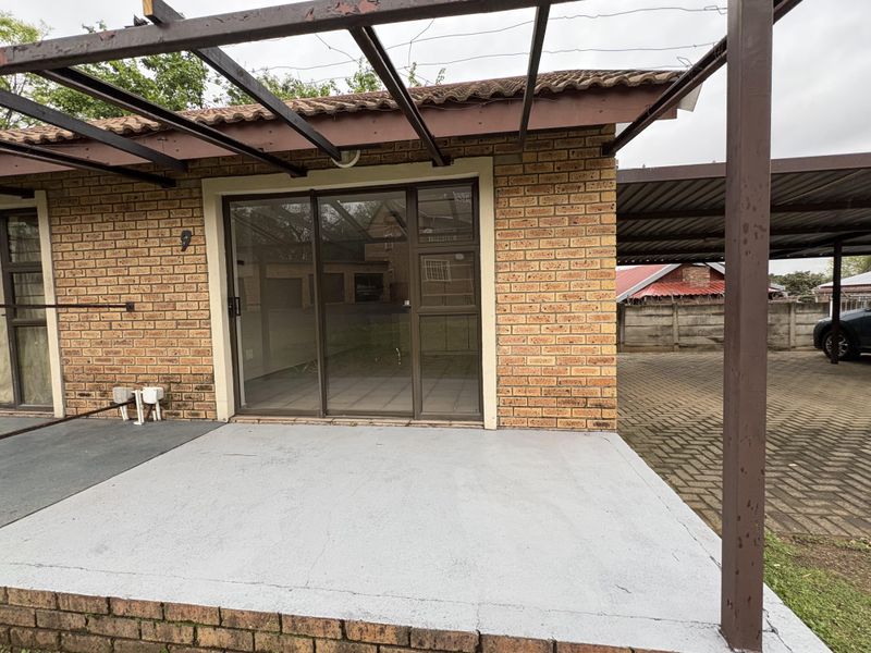 A covered patio area featuring a brick facade, sliding glass doors, and a metal pergola frame, showing signs of wear and requiring maintenance.