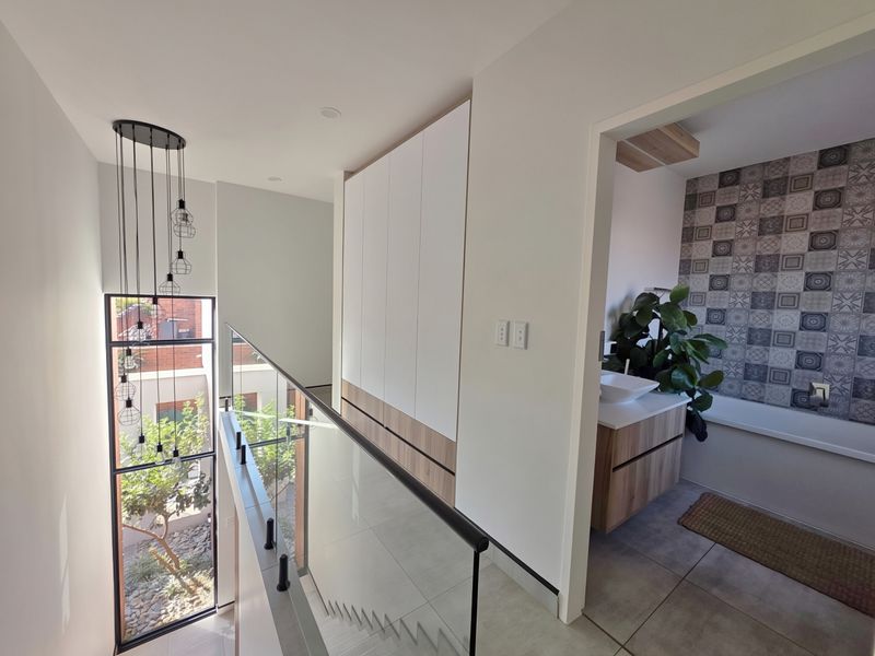 Modern upper-level landing featuring a glass balustrade, floor-to-ceiling window, built-in storage cabinetry, and contemporary pendant lighting, leading into an adjacent bathroom.