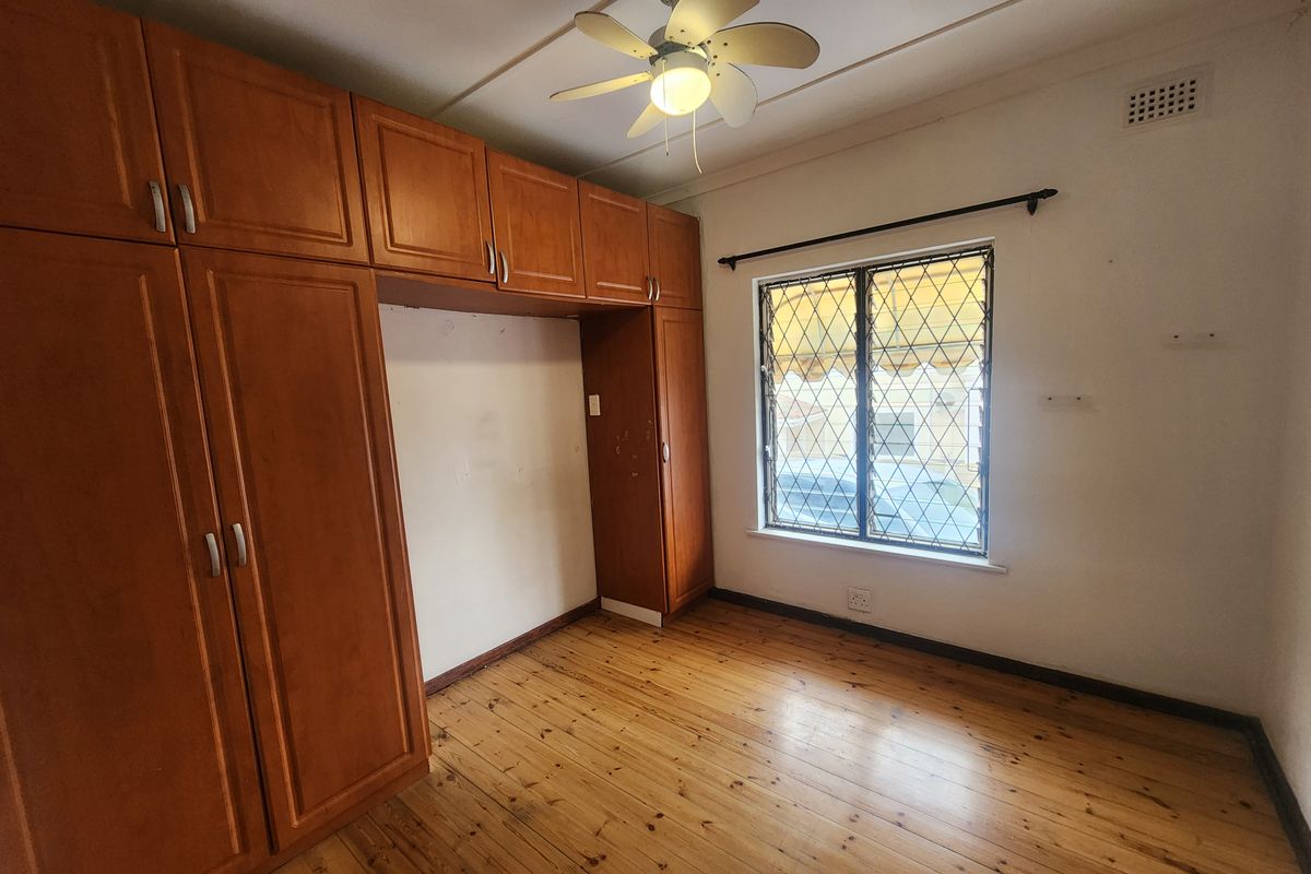 A vacant bedroom featuring timber flooring, built-in wooden cupboards, a ceiling fan, and a window with security bars.