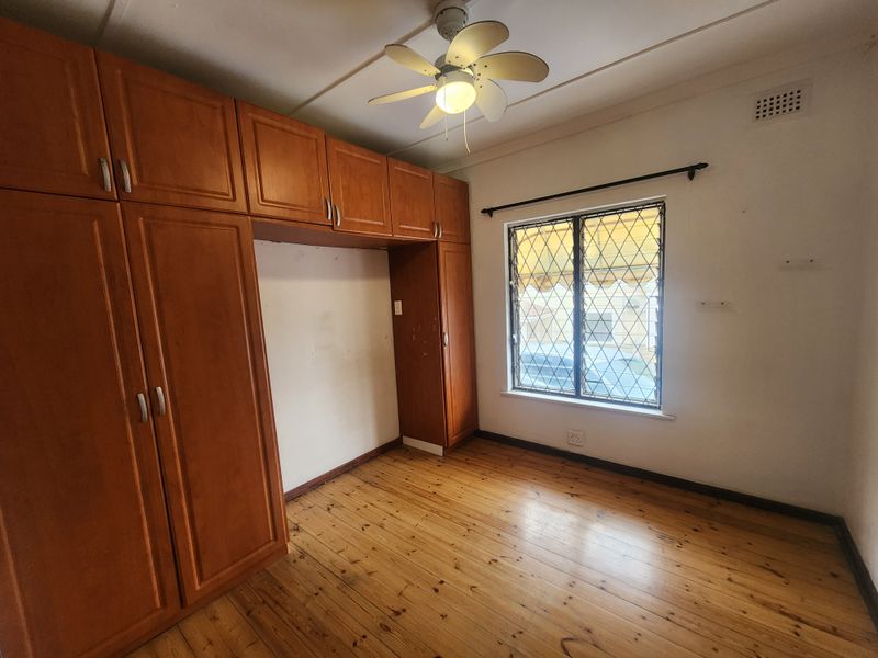 A vacant bedroom featuring timber flooring, built-in wooden cupboards, a ceiling fan, and a window with security bars.