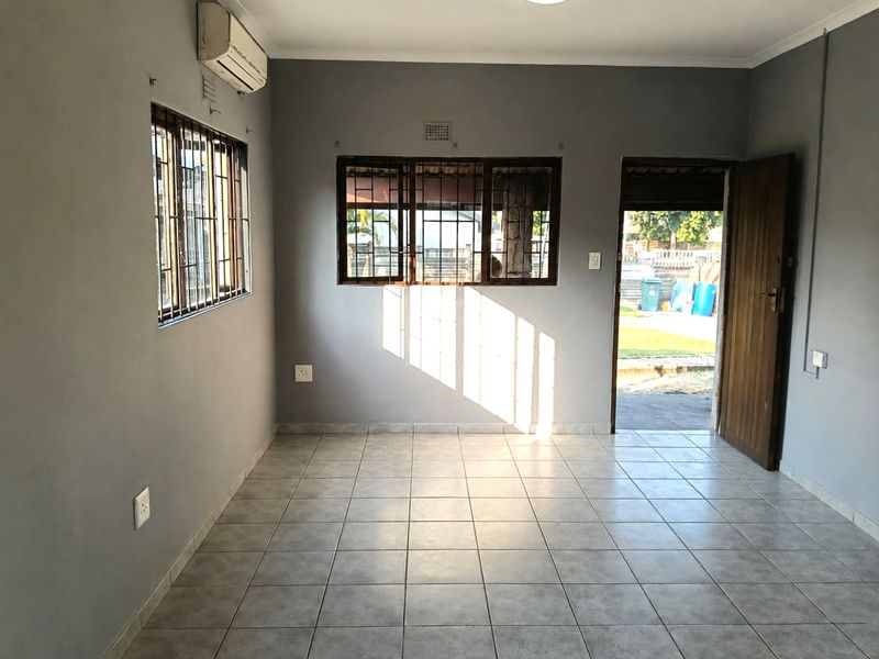 A vacant living room featuring tiled flooring, a wall-mounted air conditioning unit, and security bars on the windows with direct access to the exterior.