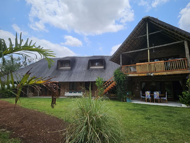 Rear view of a double-story house featuring a traditional thatch roof, brick exterior, wooden balcony, and a spacious garden area.