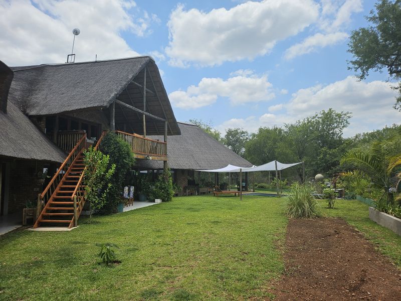 Rear view of a thatched-roof house featuring a wooden staircase, balcony, and a landscaped garden with a swimming pool and shade sail.