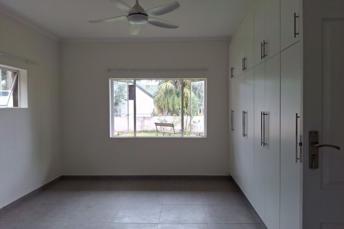 A bright, empty bedroom featuring neutral tiled flooring, a ceiling fan, and extensive white built-in cupboards for ample storage.
