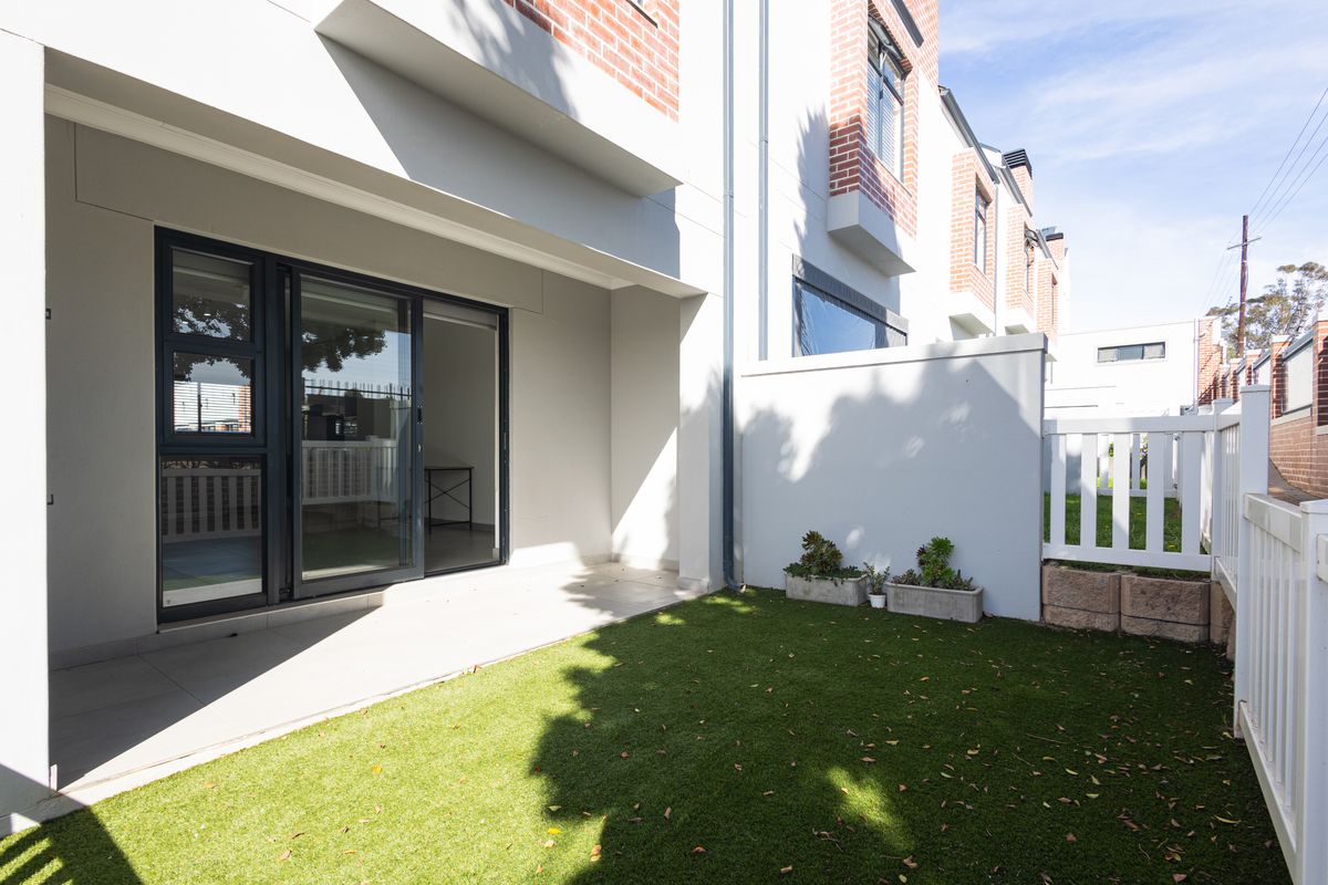 A private rear courtyard featuring a tiled patio area, artificial lawn, and secure boundary fencing, accessible via sliding glass doors.
