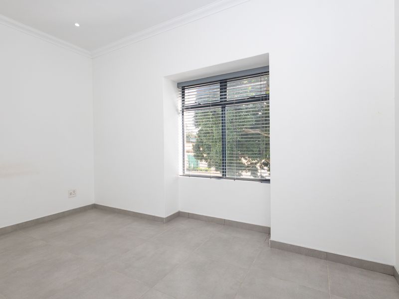 A bright, empty bedroom featuring neutral tiled flooring, clean white walls, and a window fitted with venetian blinds.