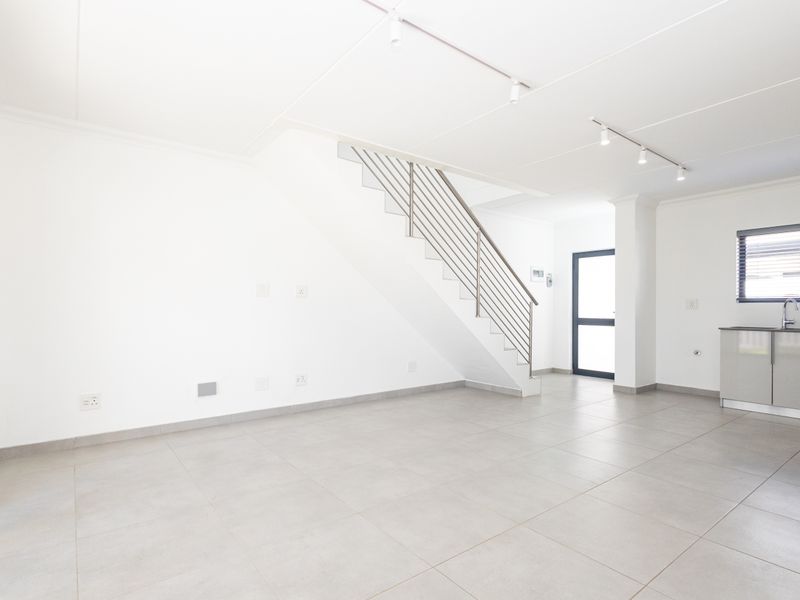 Bright open-plan living area featuring tiled floors, a modern staircase with metal balustrade, track lighting, and a compact kitchenette corner.