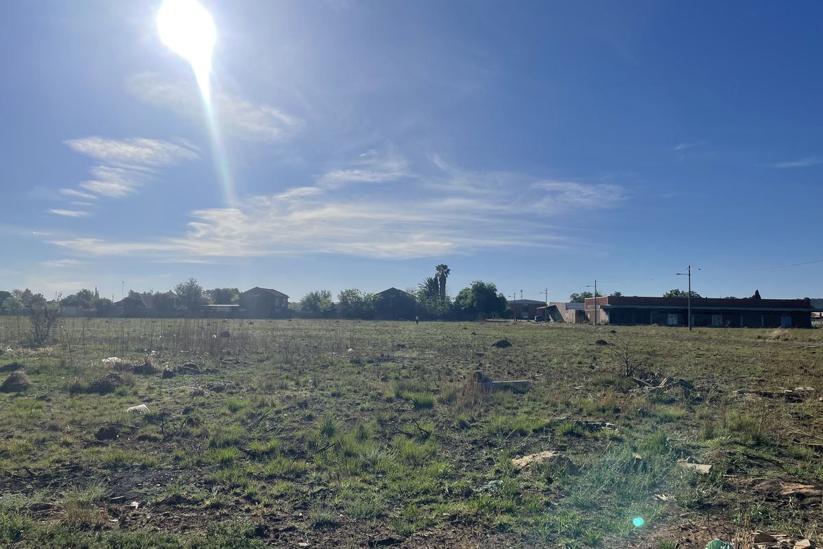 A wide-angle view of a vacant residential plot of land with natural grass cover and surrounding neighborhood structures in the distance.