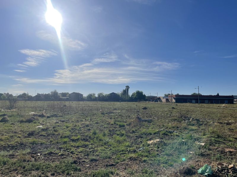 A wide-angle view of a vacant residential plot of land with natural grass cover and surrounding neighborhood structures in the distance.