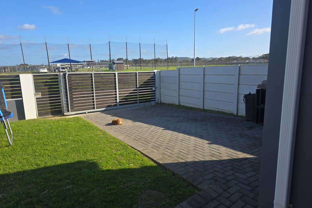 A rear exterior view featuring a paved driveway, a slatted metal gate, and a precast concrete boundary wall adjacent to a lawn area.