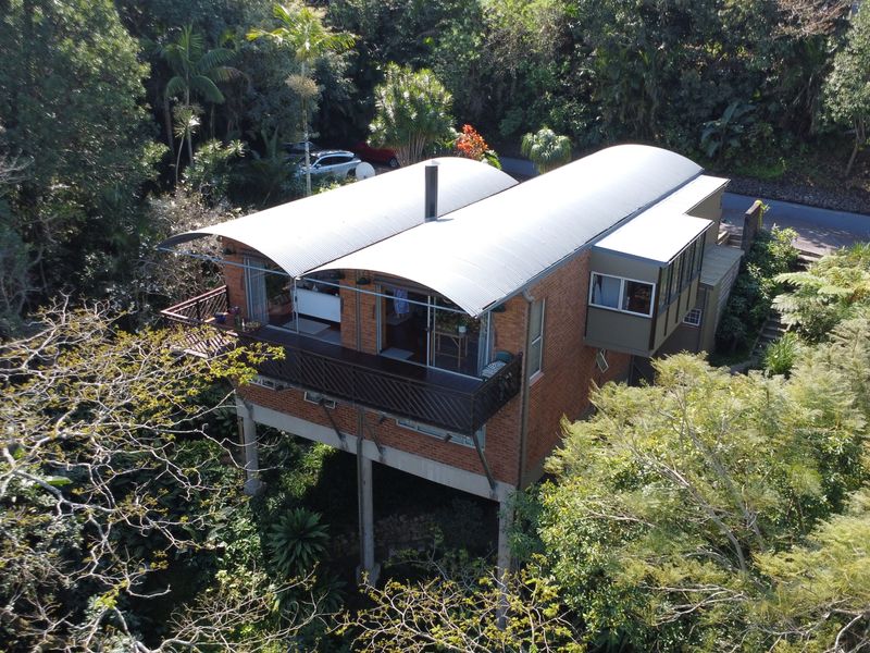 Aerial view of a unique townhouse featuring a curved metal roof, brick exterior, and elevated structure nestled within a lush, wooded environment.