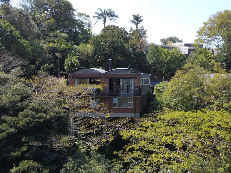 An aerial view of a multi-level brick townhouse nestled within a lush, wooded environment, featuring a distinctive curved roof and elevated balcony structure.