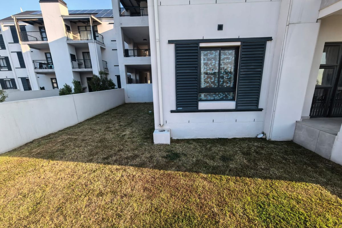 A view of the rear garden area featuring a lawn, boundary wall, and exterior window shutters on the apartment building facade.
