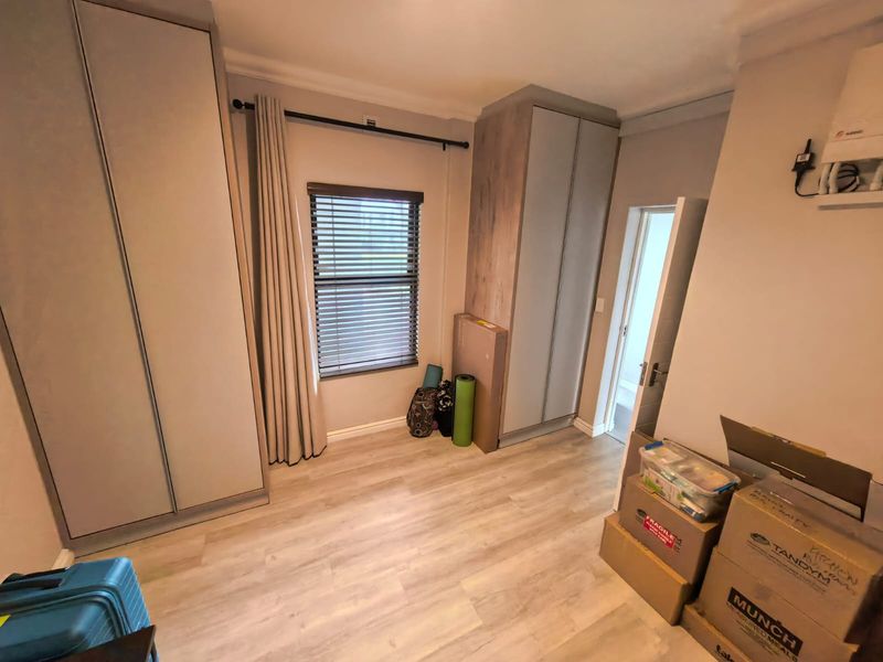 A bright bedroom featuring modern laminate flooring, neutral walls, and ample built-in cupboard storage with venetian blinds.