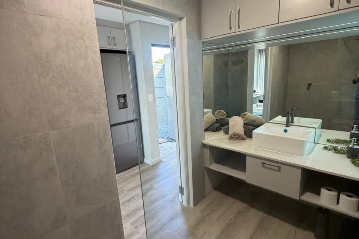Modern bathroom featuring a vessel sink, wall-mounted vanity with storage, large mirror, and neutral grey tiling, with a view into the adjacent kitchen area.