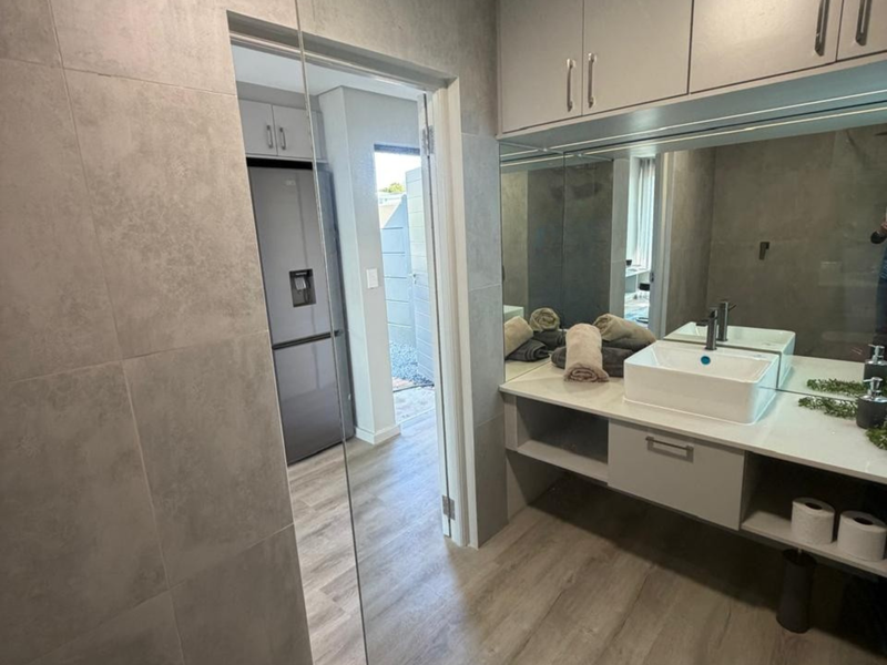 Modern bathroom featuring a vessel sink, wall-mounted vanity with storage, large mirror, and neutral grey tiling, with a view into the adjacent kitchen area.