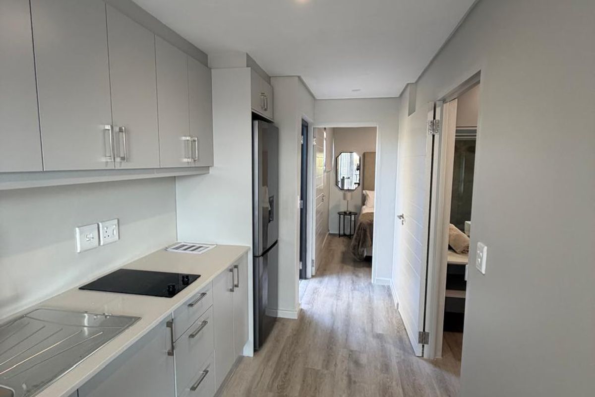 Modern galley-style kitchen featuring sleek grey cabinetry, integrated electric hob, stainless steel sink, and light wood-look laminate flooring, leading into the bedroom area.