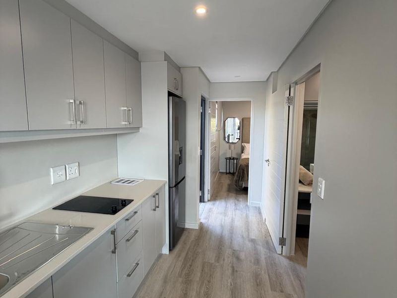 Modern galley-style kitchen featuring sleek grey cabinetry, integrated electric hob, stainless steel sink, and light wood-look laminate flooring, leading into the bedroom area.