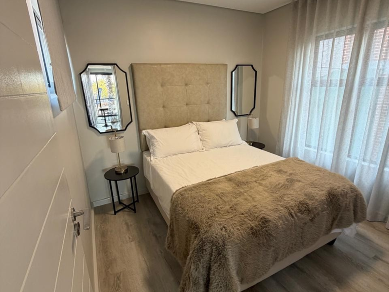 A well-maintained bedroom featuring light-toned laminate flooring, neutral wall finishes, and large windows with fitted curtain tracks.