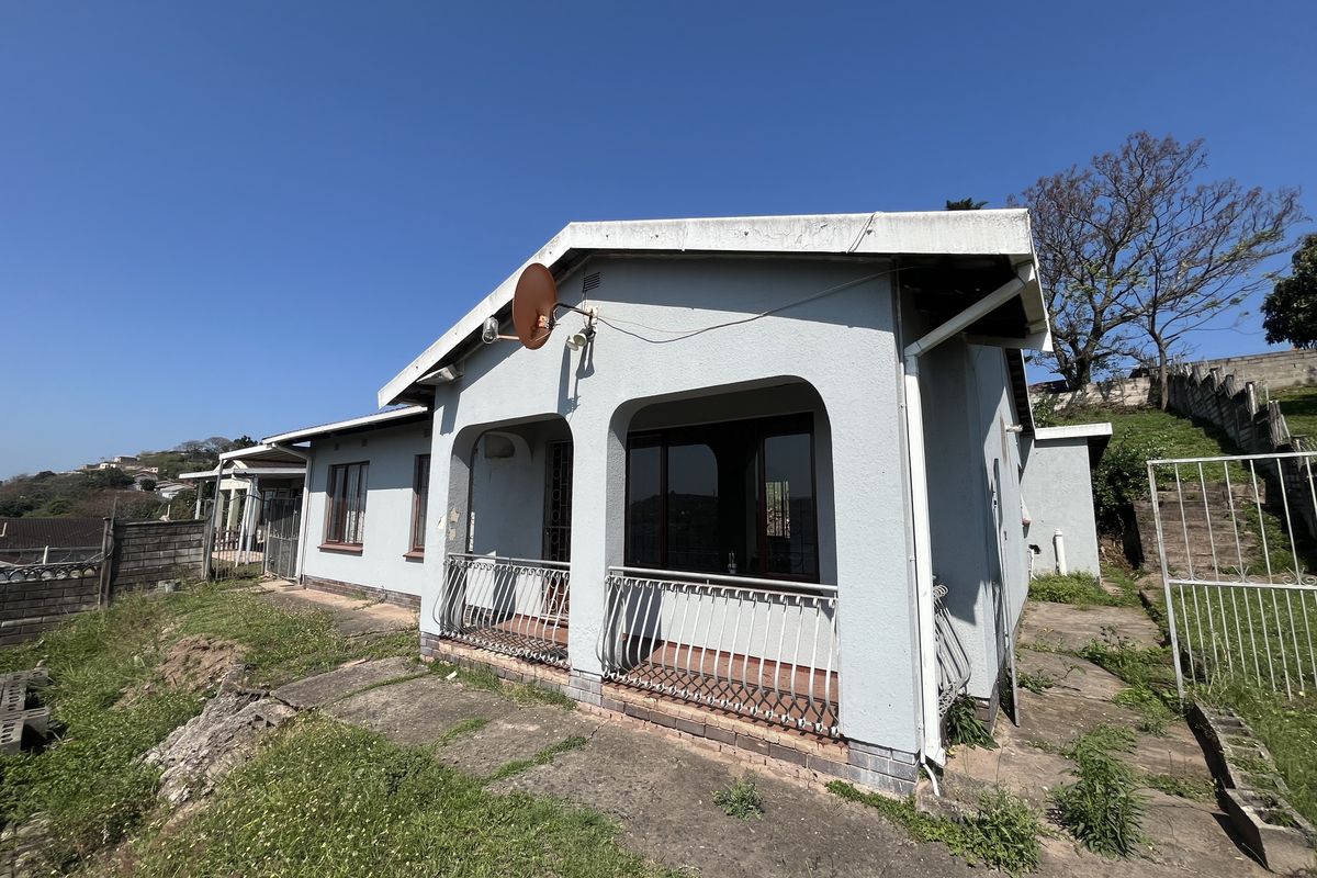 Rear exterior view of the house featuring a covered patio with metal balustrades, concrete paving, and surrounding garden area requiring maintenance.