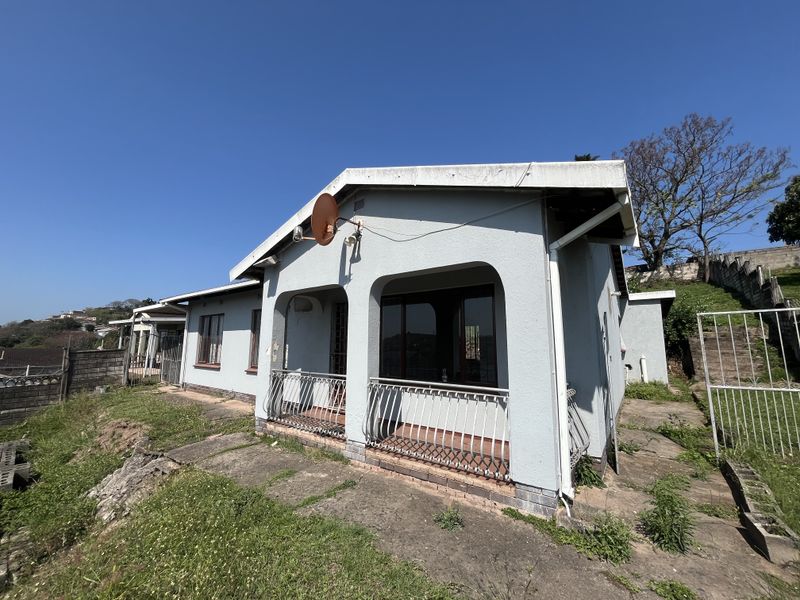 Rear exterior view of the house featuring a covered patio with metal balustrades, concrete paving, and surrounding garden area requiring maintenance.