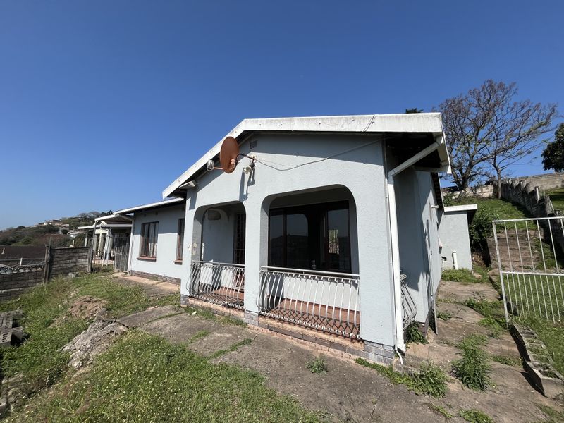 Front exterior view of a single-story house featuring a covered patio, metal balustrades, and a concrete driveway, set on a sloped plot.
