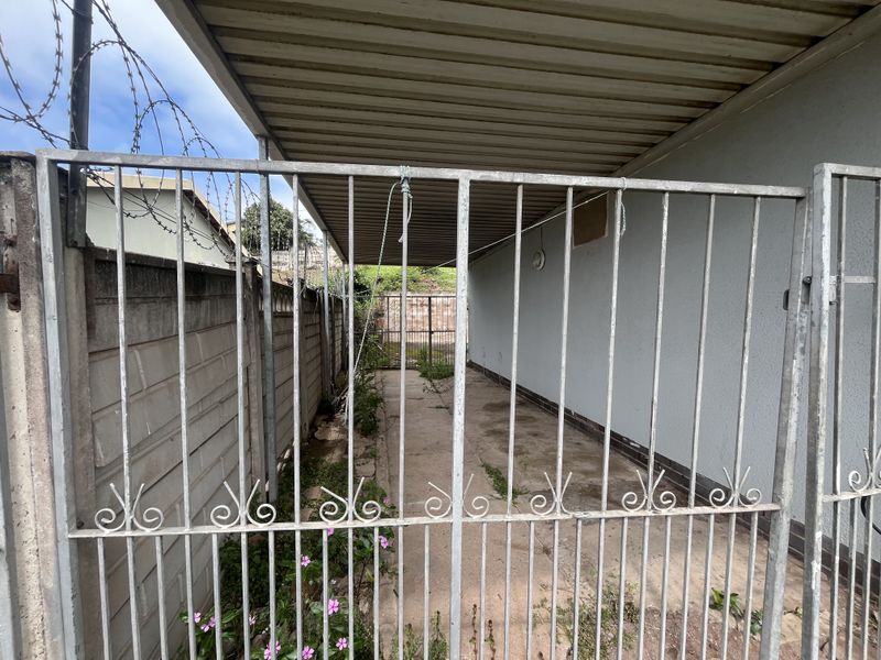 A narrow, covered outdoor side passage featuring a metal security gate, precast concrete boundary wall with razor wire, and paved flooring with visible weed growth.