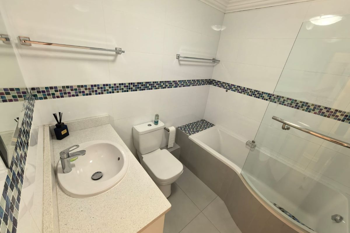 A clean, modern bathroom featuring a vanity basin, toilet, and a bathtub with an integrated glass shower screen and decorative mosaic tile accents.