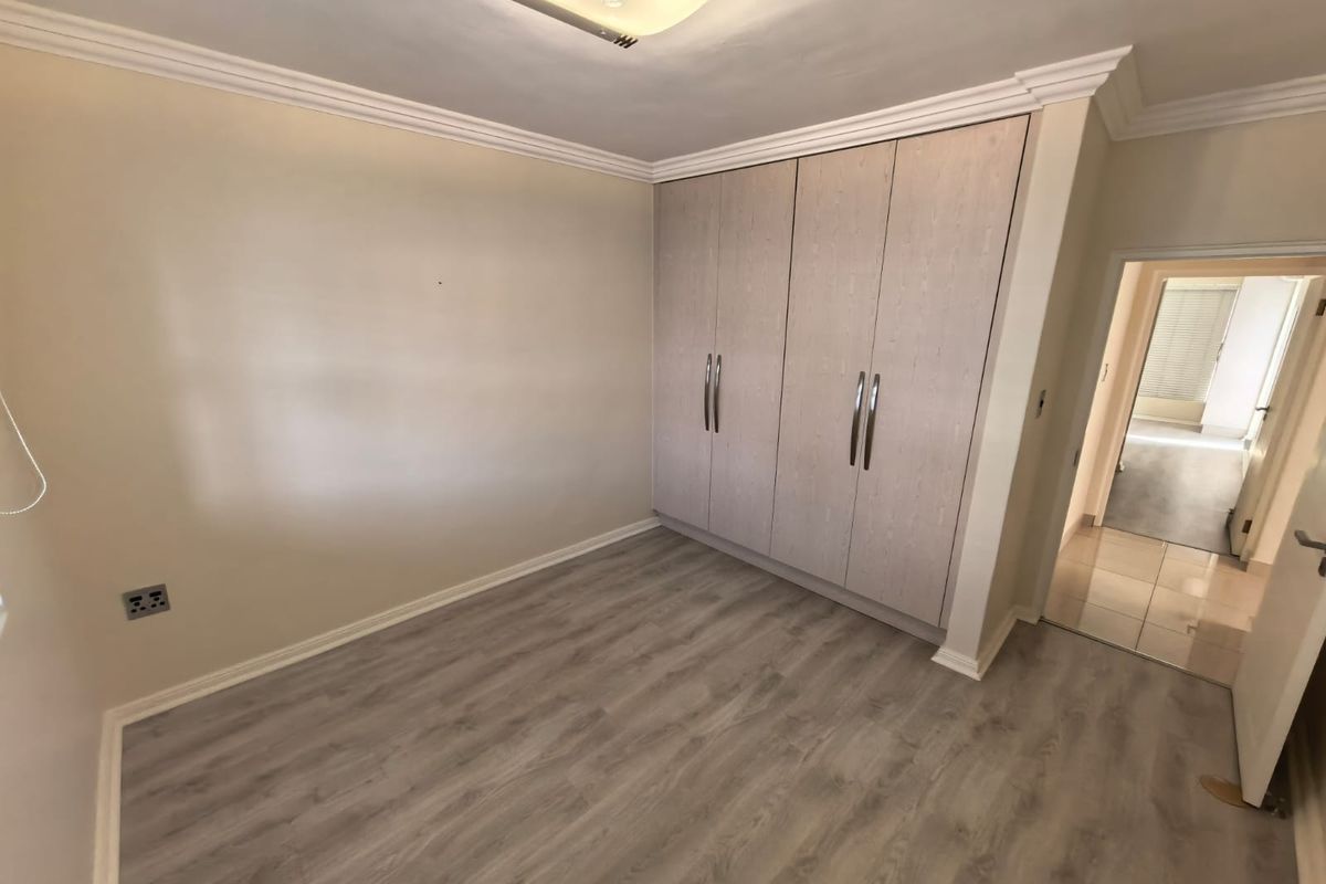 A clean, empty bedroom featuring modern light-toned laminate flooring, built-in cupboards with metallic handles, and decorative ceiling cornicing.