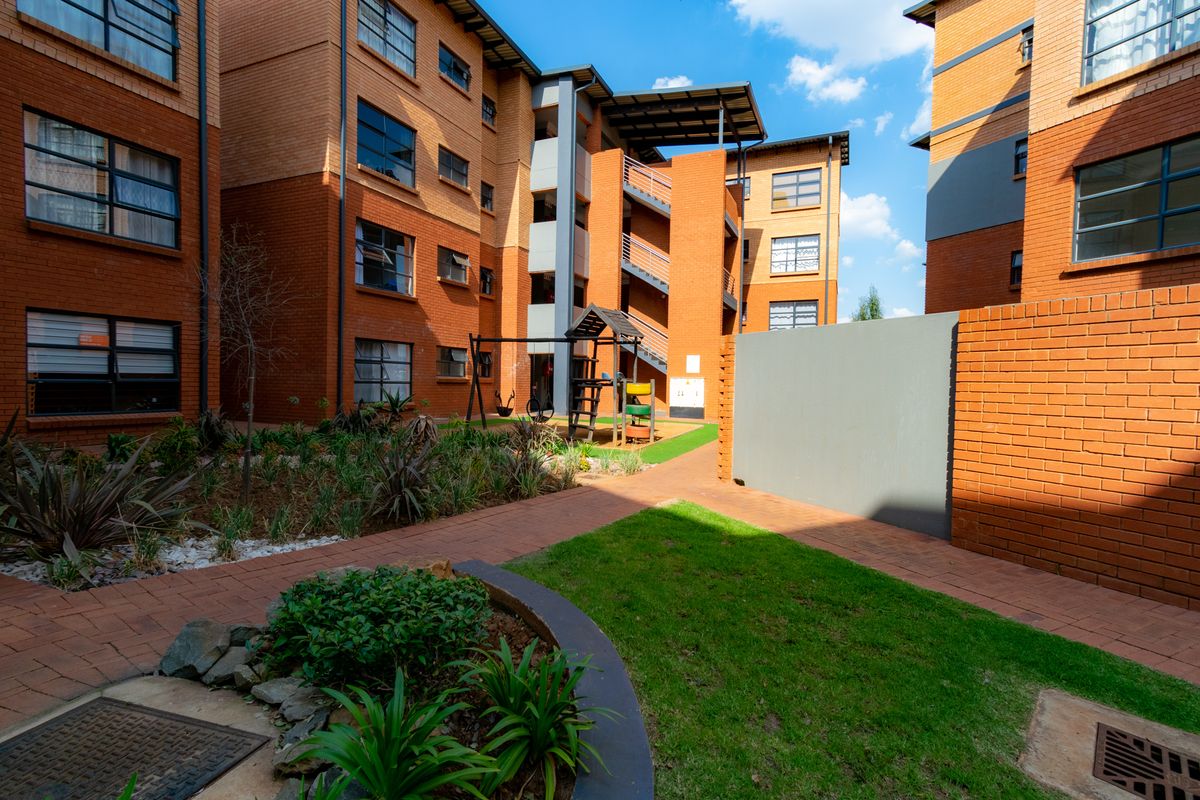 A well-maintained apartment complex courtyard featuring brick facades, paved walkways, landscaped garden beds, and an outdoor play area.