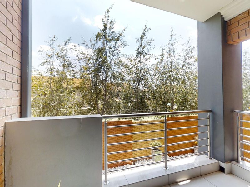 A clean, covered balcony featuring tiled flooring and a modern stainless steel balustrade, overlooking a landscaped garden area.