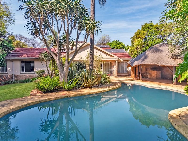 Rear garden view featuring a swimming pool, a thatched lapa entertainment area, and a well-maintained home exterior with solar panels.