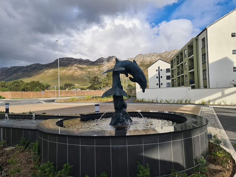 A well-maintained apartment complex entrance featuring a decorative dolphin fountain, paved driveway, and scenic mountain views in the background.