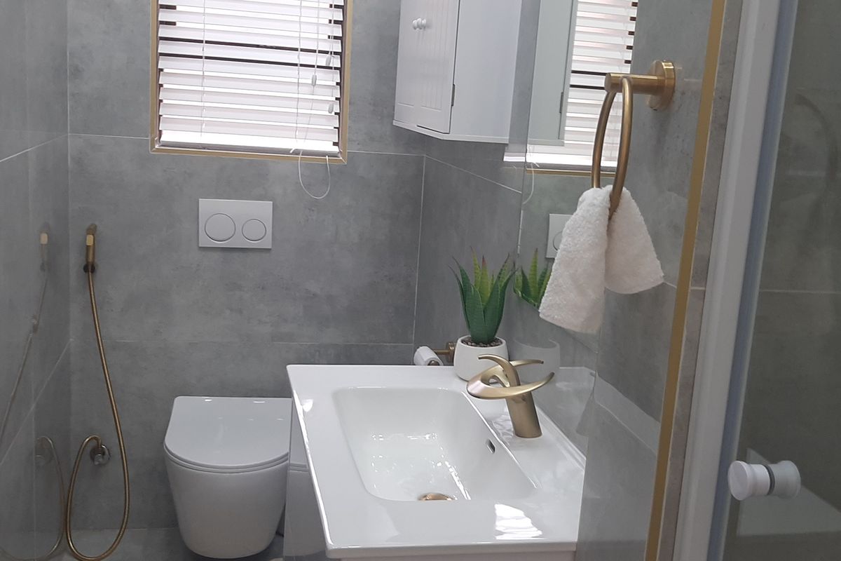 Modern bathroom featuring grey wall tiles, a wall-hung toilet with concealed cistern, vanity basin with gold-tone fixtures, and a wall-mounted storage cabinet.