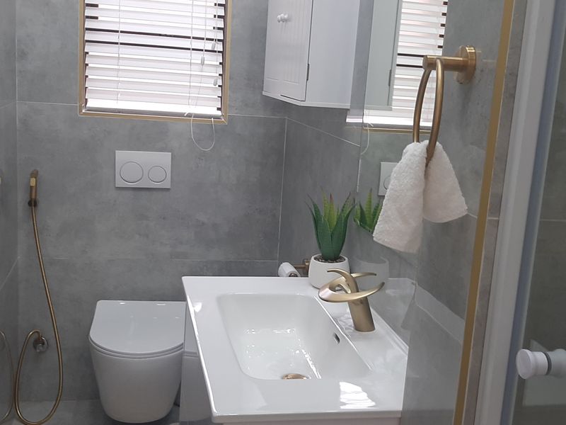 Modern bathroom featuring grey wall tiles, a wall-hung toilet with concealed cistern, vanity basin with gold-tone fixtures, and a wall-mounted storage cabinet.