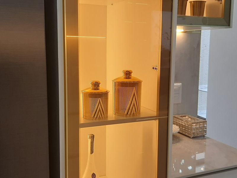 A modern kitchen detail featuring a sleek built-in display cabinet with integrated warm lighting, stone countertops, and a stainless steel refrigerator.