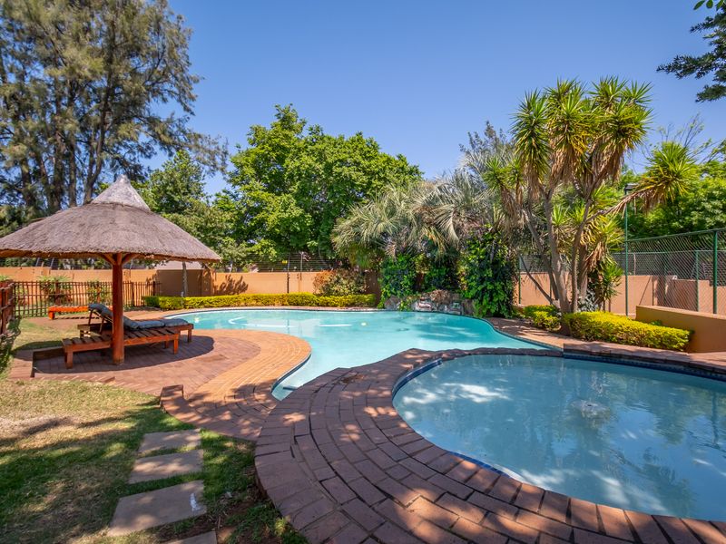 A well-maintained outdoor pool area featuring a swimming pool, a smaller circular pool, paved surrounds, and a thatched umbrella structure.