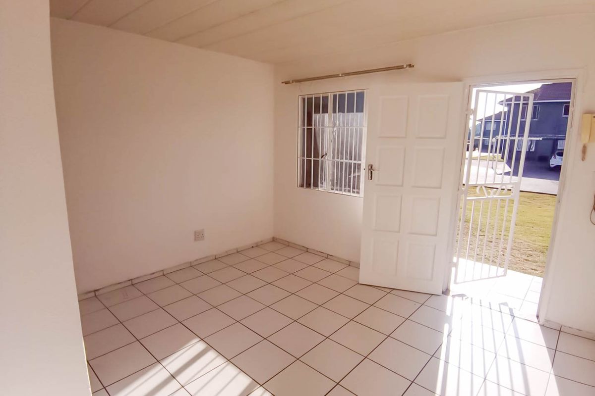 An empty, sunlit living area featuring white tiled flooring, a security gate, and direct access to the exterior.