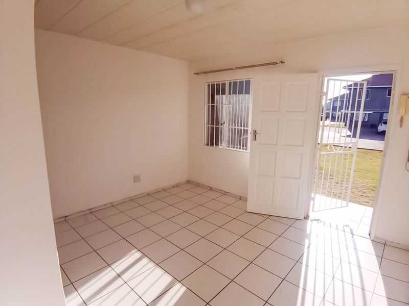 An empty, sunlit living area featuring white tiled flooring, a security gate, and direct access to the exterior.